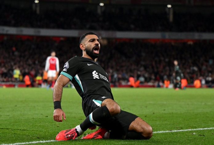 Liverpool Mohamed Salah sliding on his knees in celebration after scoring a goal, wearing a black Liverpool kit during a match against Arsenal.